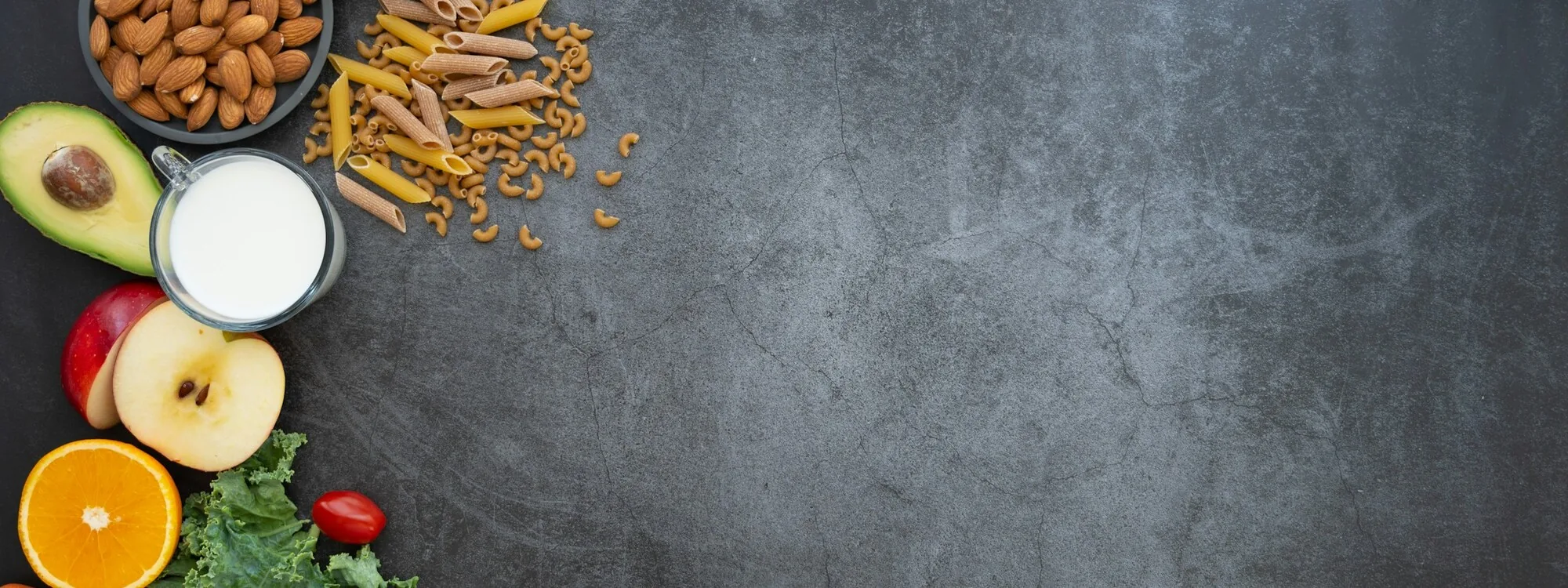 Fresh fruit, vegetables, nuts, wholegrain pasta and milk arranged on a dark slate surface